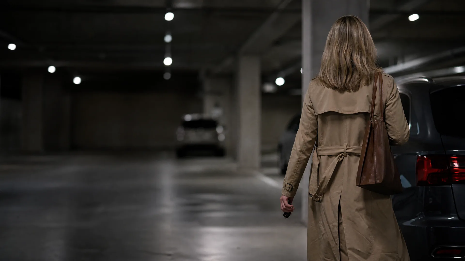 A woman in a tan trench coat with a brown shoulder bag walks away from the camera through a dimly lit underground parking garage with parked cars and ceiling lights.