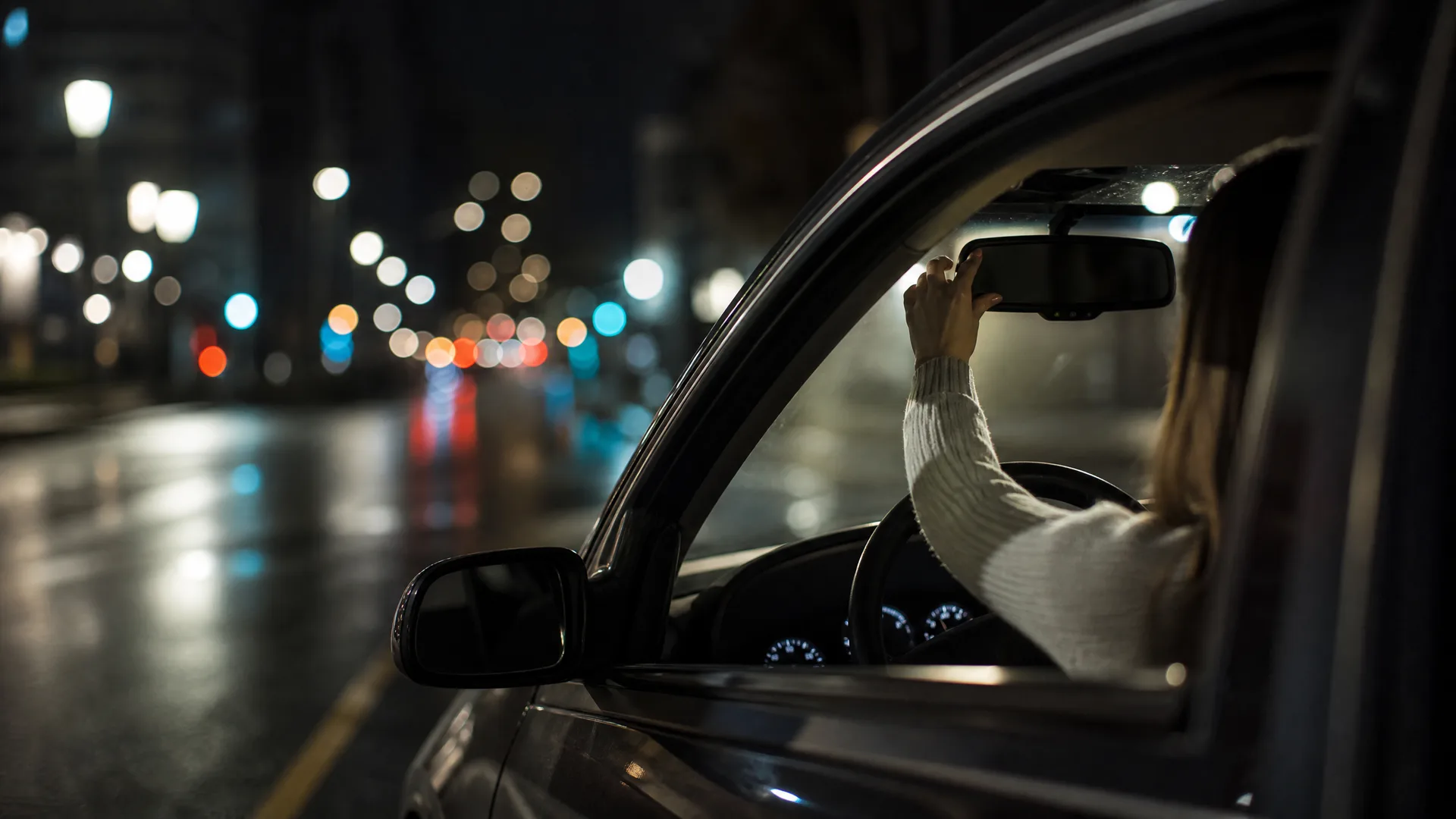 A person in a white sweater sits in a car's driver seat at night, reaching up to adjust the rearview mirror, with blurred street lights on a wet road in the background.