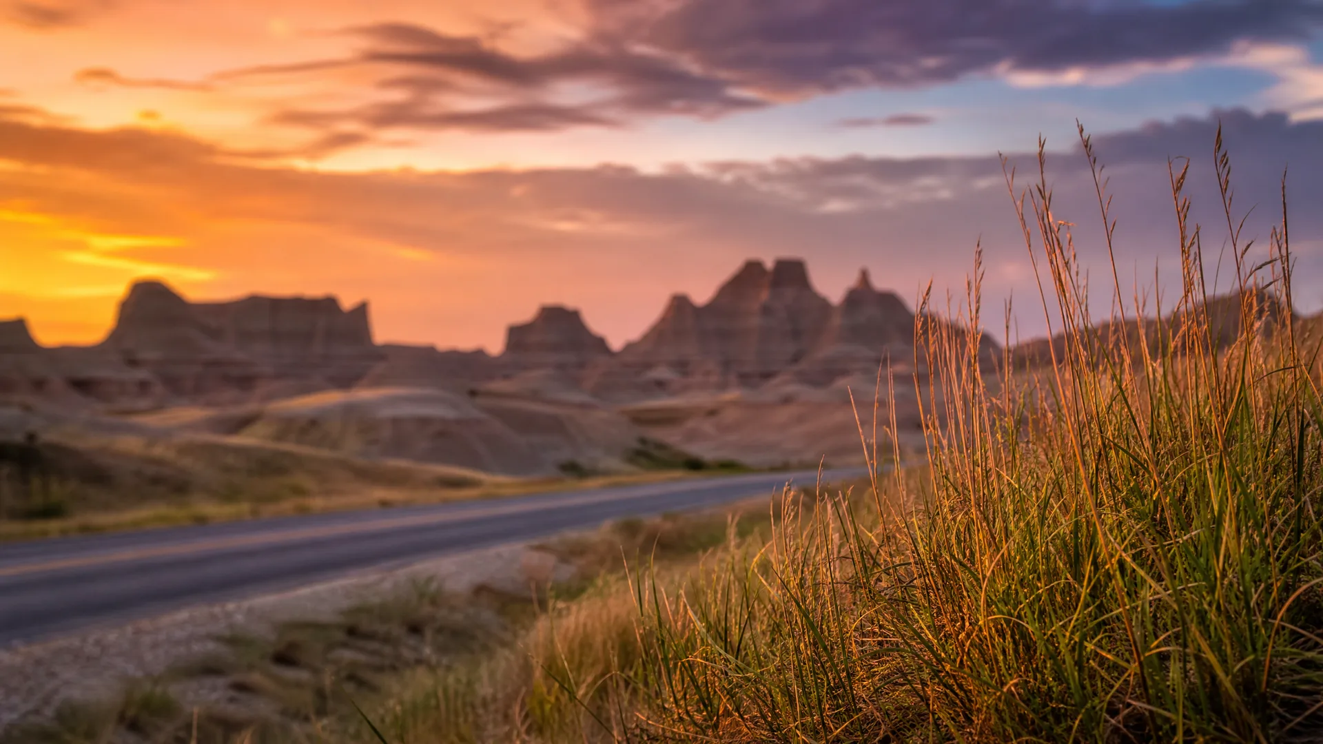 A paved road curves through dry grassland toward blurred eroded rock formations under an orange and purple sunset sky, with tall grasses in sharp focus in the right foreground.