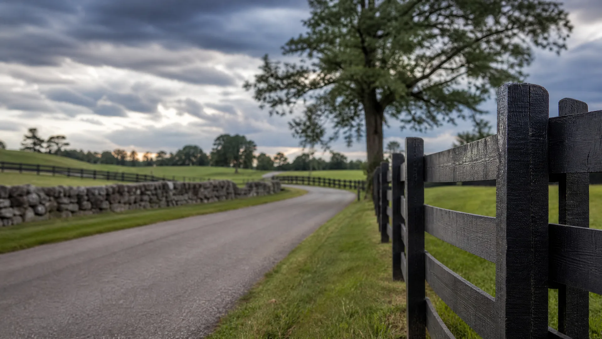 A narrow paved road curves between a dark wooden plank fence in the foreground right and a dry stone wall on the left, with grass fields, a large tree, and a cloudy sky.